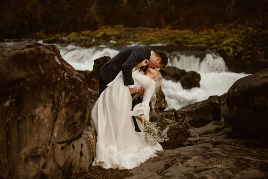 HD mountain waterfall river photo front view of newly married couple in formal bridal white dress blond long hair bride holding flower bouquet and man in black suit bending his wife on her back during french kissing her lips in thick forest by a expert wedding photographer standing on a rock at evening.