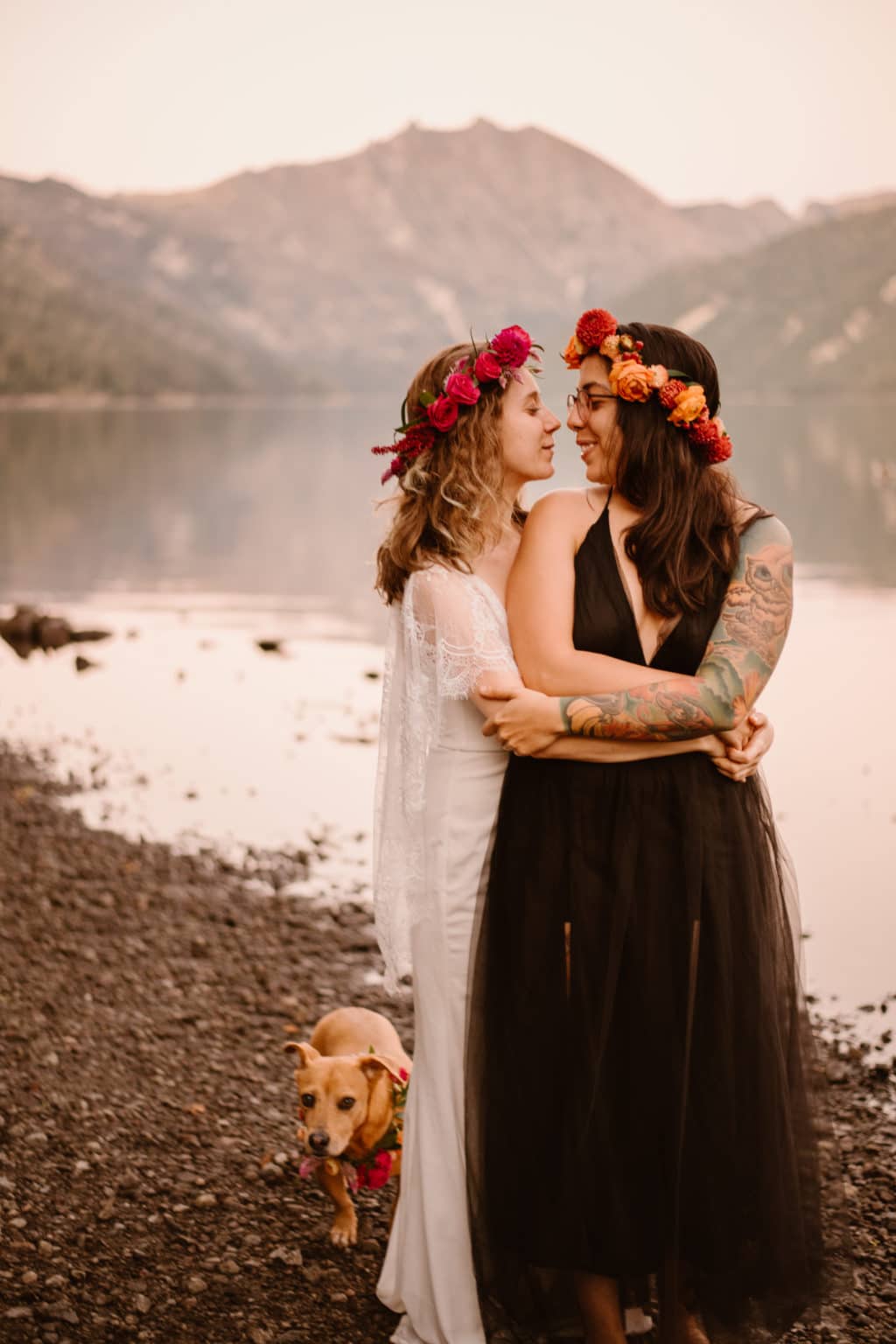 Perfect gay marriage hugging playing images of two tatooed woman near Mount Saint Helens lake in roman long black skirt and white bridal dress with floral headband crown and their brown dog by a talented wedding photographer during summer in July.
